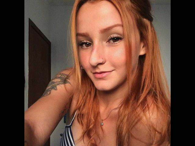 Ruivaxx with long red hair, small nose ring, and tattoo on left arm, wearing striped top, smiling softly, showing small tits, and cute face.