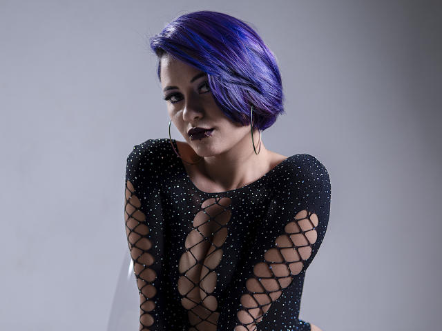 Cassie Jones met een paars kort kapsel, een zwarte kanten top die haar borsten laat zien, donkere lipstick en oorringen, kijkt naar de zijkant, met haar handen in de buurt van haar borst..