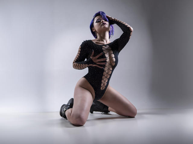 CassieJones with purple hair, kneeling, wearing a black, fishnet, cut-out bodysuit, showing her tits, with one hand on her chest and the other on her head, in boots.
