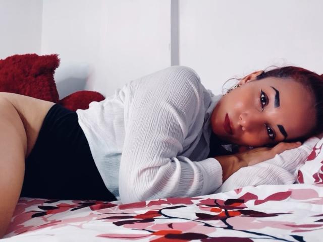 AmberDemons lies on a bed, wearing a white ribbed shirt and black high-waisted panties, showing her bare ass. She has red lipstick, arched eyebrows, and a red pillow.