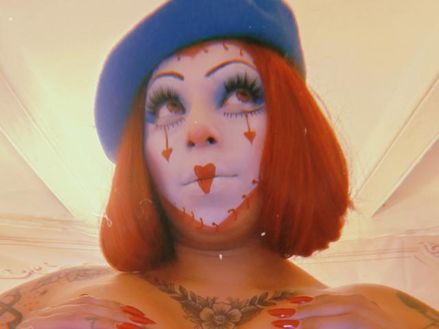 Chili, solo milf with red bob, blue beret, face painted with stitched heart, tongue out, tattoos on chest, looking up.