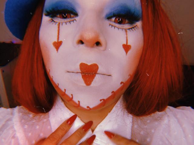 Chili is performing cunnilingus facepaint on herself, with blue eyeshadow, red lipstick, heart-shaped makeup, and red nails touching her chin.