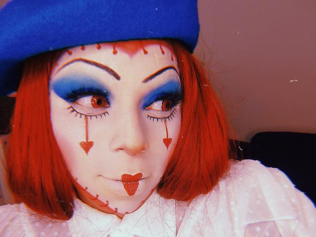 Chili con el pelo rojo está usando un sombrero azul, su cara pintada con sombra de ojos azul, corazones de sangre y lápiz labial, mirando intensamente.