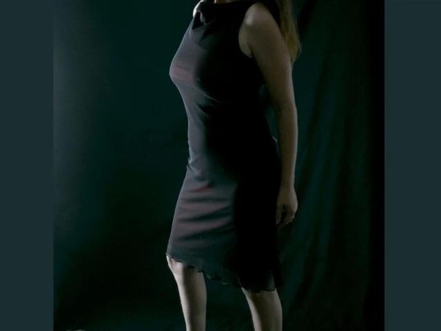 monelive-hot standing, wearing a tight black dress, showing a small, subtle, red stain near the ass, dark, shadowy background, dress is sleeveless, hands by her sides.