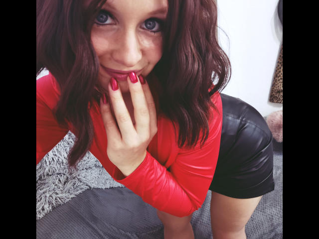 Annekehot-sex with red nails, red top, and black leather skirt, bending forward, covering her mouth with her hand, and smiling at the camera.
