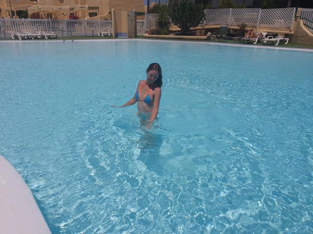 Sweet-nina in a blue bikini, sitting in a pool with clear water, her tits above the surface, long dark hair, smiling, white fence in background, sunny day.