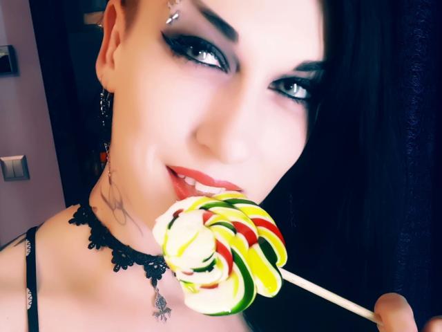 Evelinka, séduisante avec ses cheveux foncés, son maquillage gothique et son collier en dentelle, tient une sucette colorée qu'elle porte contre ses lèvres rouges, et on peut apercevoir un tatouage sur son cou..