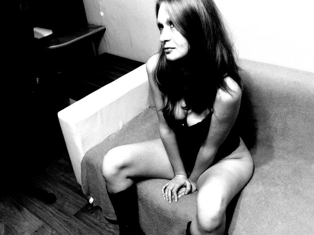 Black-and-white photo of SweetElisa1-hot sitting on a couch, legs apart, wearing a dark bodysuit that shows her tits, with her hands on her thighs and looking sideways.
