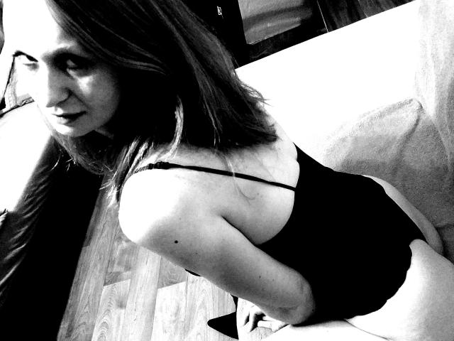Black-and-white photo of SweetElisa1-hot with long hair, wearing a spaghetti-strap black top, sitting on a couch, showing her back and looking over her shoulder, ass visible.