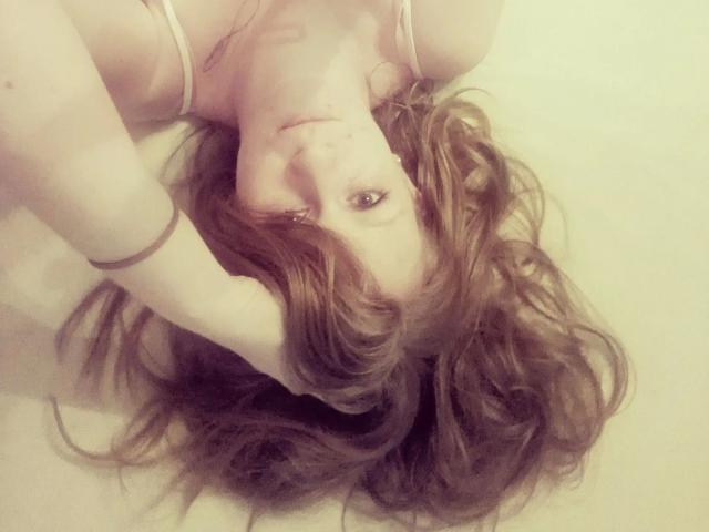 SweetElisa1-hot lying on her back with wavy brown hair, her hand in her hair, wearing a white bra, looking up with a slight smile, soft light.