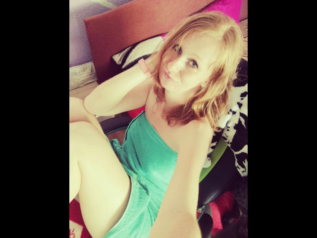 SweetElisa1-hot with blonde hair, wearing a green towel dress, sitting with legs up, hand on cheek, looking at camera, soft smile, pale skin, slight wavy hair, casual bedroom.