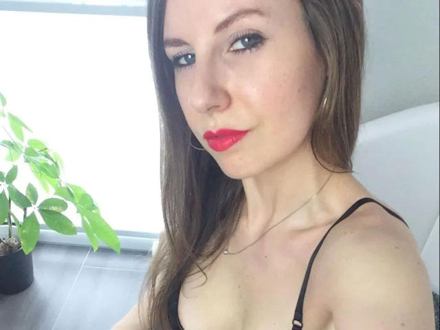 MaryJane-X with blue eyes, red lipstick, and brown hair, wearing a black spaghetti strap top, showing her tits. Potted plant on left, bright window behind.