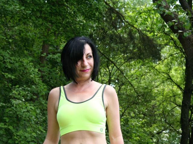 Inga-Star with short black hair, wearing a neon green sports bra, showing small tits, standing in a forest, smiling slightly, looking to the side.