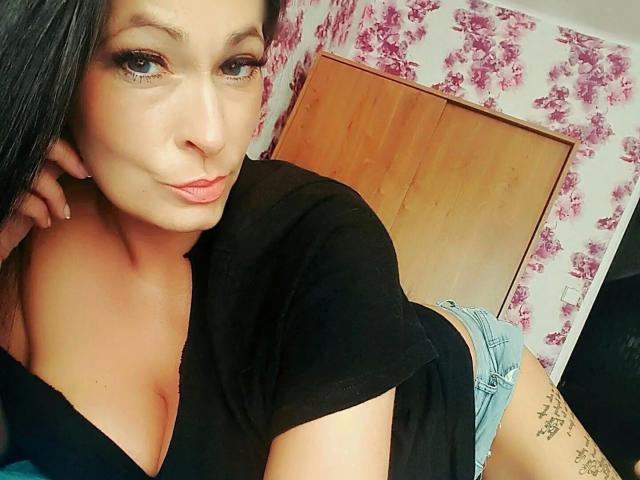 TaliaVaiolet-hot with long black hair, blue eyes, and pink lips, wearing a black top and blue shorts, showing her tits and tattoo on her right thigh.