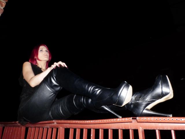 FetishQueenofPain-hot with red hair, black sleeveless top, and thigh-high black boots with high heels, sitting on a red bench, legs up, against a dark background.