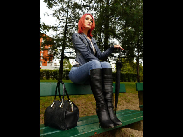 FetishQueenofPain-hot with red hair, blue jacket, and tight jeans sits on a green bench, wearing black knee boots, holding a black purse, in a forested area.