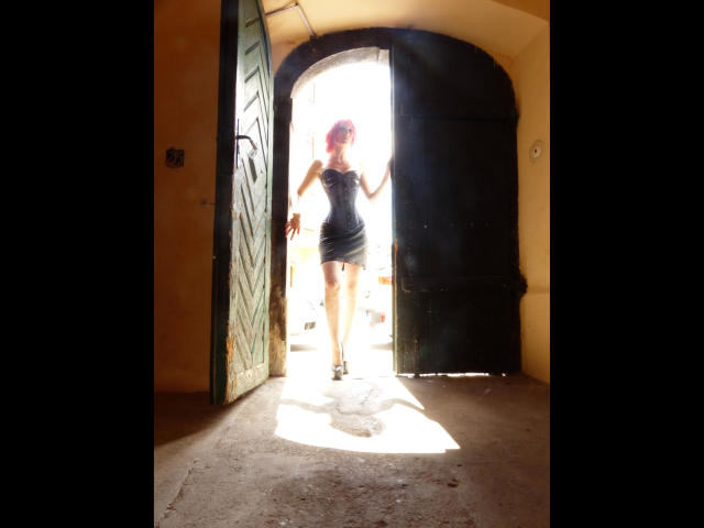 FetishQueenofPain-hot with red hair, wearing a tight black dress and heels, standing in a sunlit doorway, her hands on the door, casting a shadow on the floor.