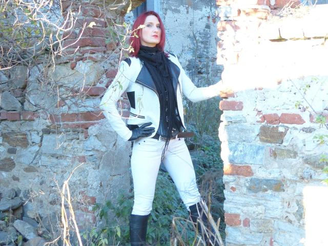 FetishQueenofPain-hot with red hair, white jacket, black scarf, white pants, black boots, and black gloves, standing against a brick and stone wall. Hands on hips, looking up.
