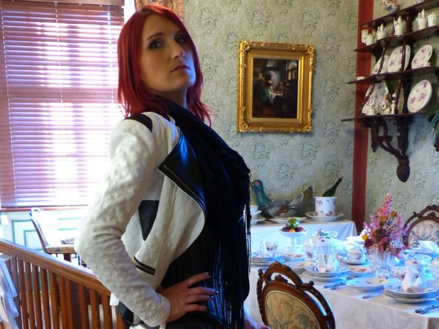 FetishQueenofPain-hot with red hair, wearing a white jacket and black dress, standing in a room with a table set for tea, hands on hips, looking at camera.