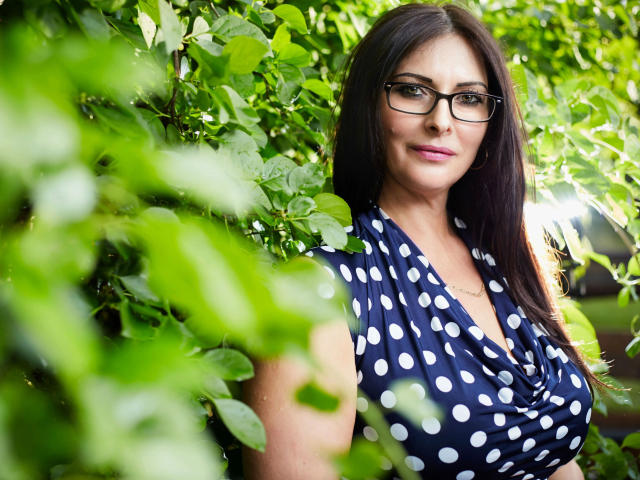 SophieSexxy, très séduisante avec ses cheveux bruns foncés, ses lunettes et sa robe bleue à pois, dévoilant ses grandes seins, se tient debout dans un feuillage vert. Elle sourit discrètement et a l'air adorable..