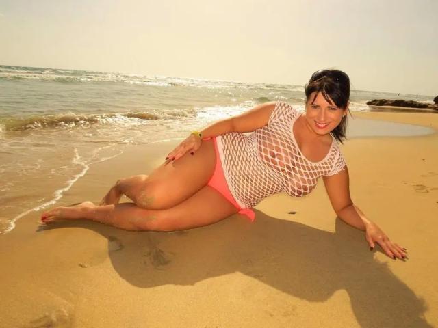 LaLeticia liegt auf einem sandigen Strand, trägt ein weißes Netzoberteil und pinke Bikini-Hosen, ihre Brüste sind durch das Netz sichtbar, sie lächelt in die Kamera, im Sonnenlicht, mit Wellen im Hintergrund..