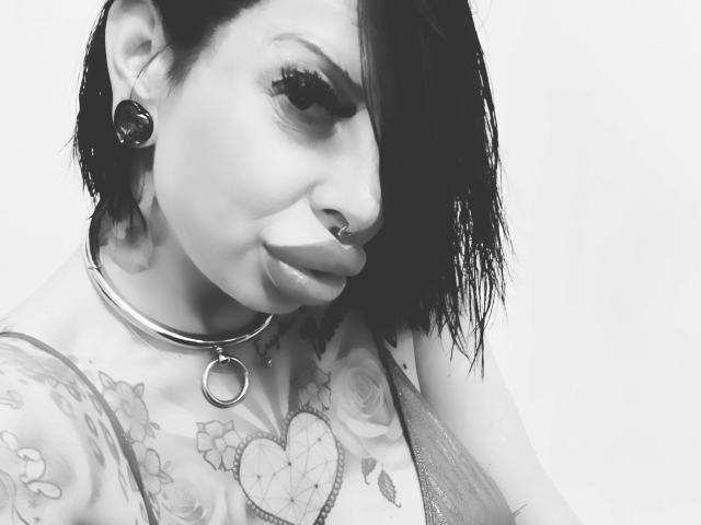 NinaVegas-hot with black hair, nose ring, and ear gauges, wearing a choker with a ring, showing tattooed tits and a heart on her chest. Black-and-white photo.