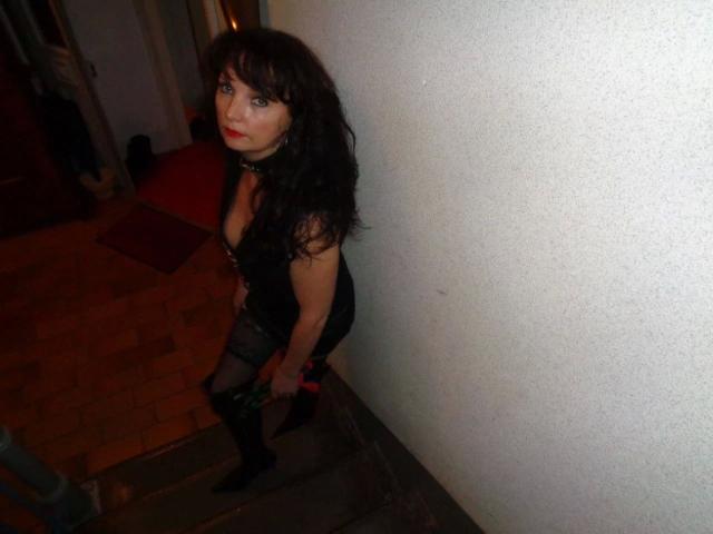 Hot-Lady34 with long dark hair, black dress, and sheer stockings stands on wooden stairs, looking up with red lipstick, wearing black boots, and a black choker.