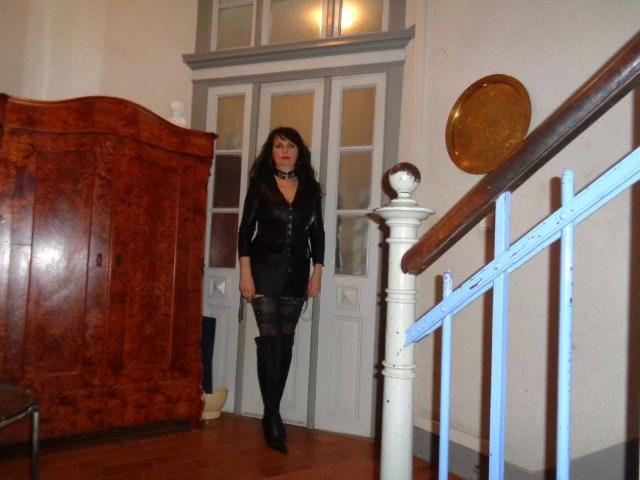 Hot-Lady34 stands in a room wearing a black dress, lace stockings, and boots, with long curly hair, and a confident expression. She's next to a wooden staircase.
