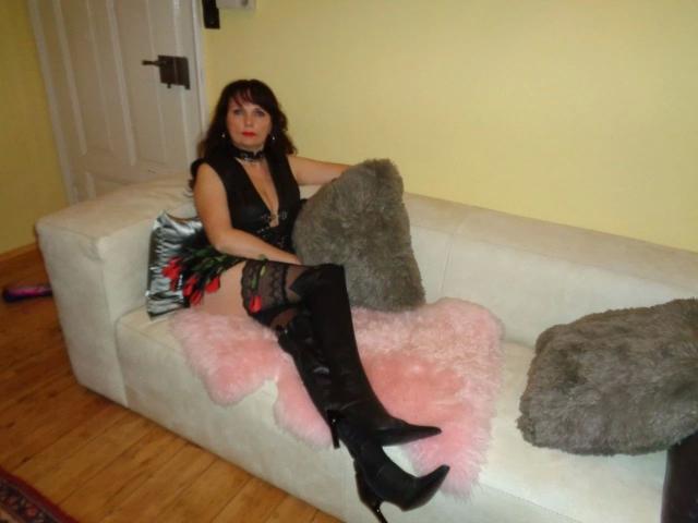 Hot-Lady34 with black lace lingerie, thigh-high black boots, and red rose in hand, lounging on white couch with pink and gray pillows, wooden floor, and yellow wall.