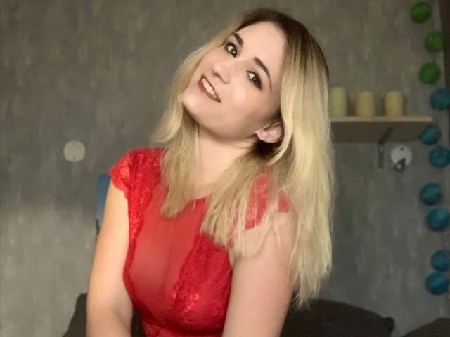 Melina-May with blonde hair, red lace top, smiling, head tilted, showing her tits, in a casual indoor setting, looking cute.