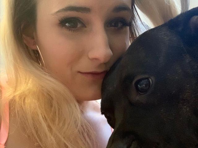 Melina-May with blonde hair, hoop earrings, and eyeliner, snuggling a black dog with shiny eyes. Her pink top is visible. She's smiling softly.