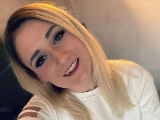 Melina-May with blonde hair, white shirt, dark eye makeup, and red lipstick, smiling at the camera. Her tits are not visible. She has a cute, sexy look.