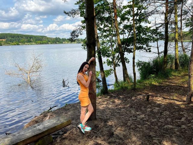 Trixie, vestida de naranja, apoyada en un árbol, mostrando su trasero, con zapatillas azul verdosas, junto a un lago, rodeada de árboles y un banco de madera..
