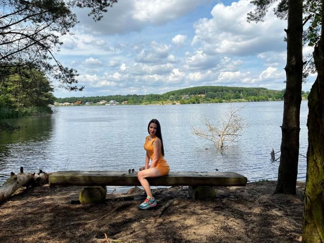 Ein Trixie-Hund, mit einem orangefarbenen Kleid und Turnschuhen, sitzt auf einem Holzstamm am See, die Arme auf den Knien, blickt in die Kamera, umgeben von Bäumen, bewölkter Himmel, Wasser im Hintergrund..