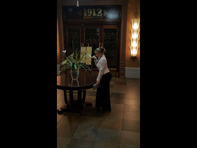 HottieKattie: sesso con il sedere rivolto verso il basso, piegata, mentre pulisce un tavolo rotondo, indossando una camicia bianca e una gonna nera, in una stanza poco illuminata con un vaso di fiori bianchi..