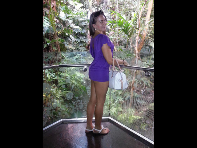 HottieKattie-sessuale, in piedi su un balcone, indossa un vestito viola, tiene una borsa bianca, mostra il sedere, pelle abbronzata, capelli scuri, sorridente, sandali bianchi, vegetazione sullo sfondo..