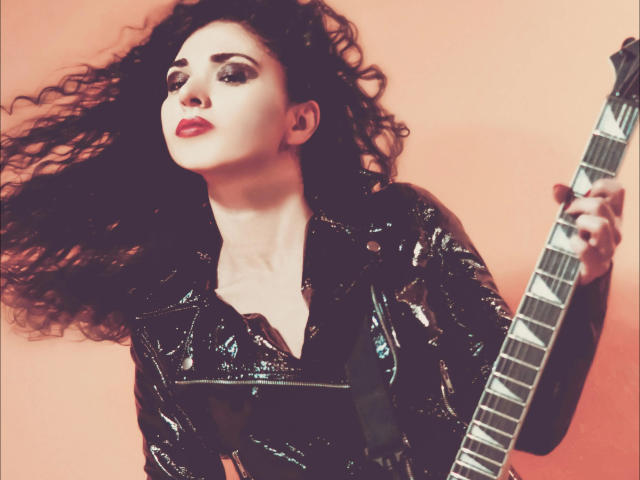 Con i capelli neri ricci, un aspetto sensuale, una giacca di pelle nera lucida e rossetto rosso, tiene una chitarra basso, guardando verso l'alto con un'espressione sicura e leggermente seducente..