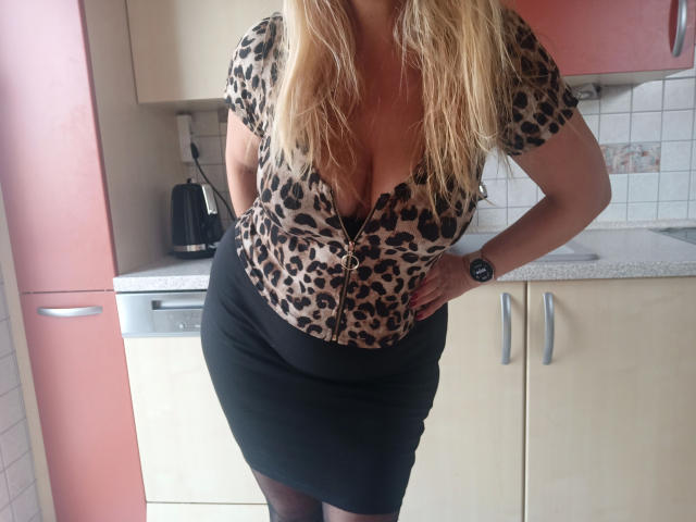 Femme aux cheveux blonds, portant un haut léopard qui met en valeur sa poitrine généreuse, une jupe noire, une montre noire, debout dans une cuisine, une main sur la hanche, et portant des bas noirs..