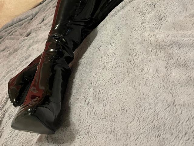 HotLeyla86-sex's shiny black thigh-high boots with a glossy finish, resting on a gray, textured blanket. Boots are positioned in the bottom left corner of the image.