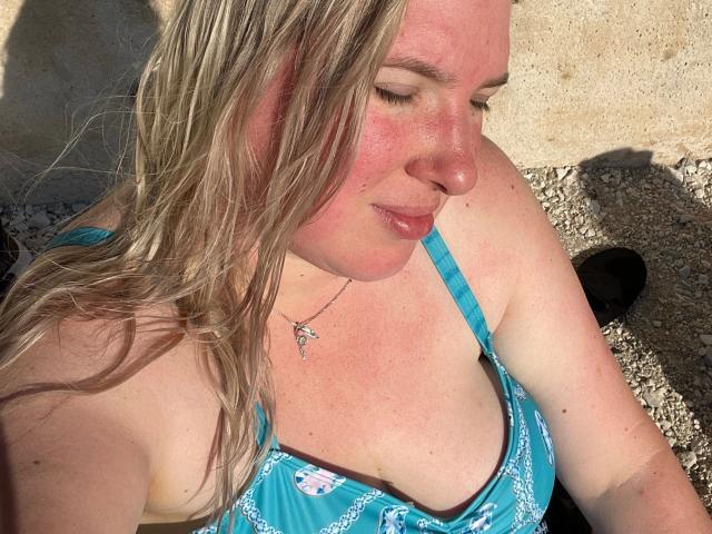HotLeyla86-sex with sunburned face, blonde wet hair, blue patterned top, small tits, pink lips, slight smile, necklace with cross, sitting in sunlight, shadow on right.