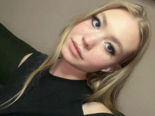 BrandyHelga-hot has blonde hair, blue eyes, and freckles on her face. She's wearing a black top, with white earbuds in. Her lips are slightly parted.
