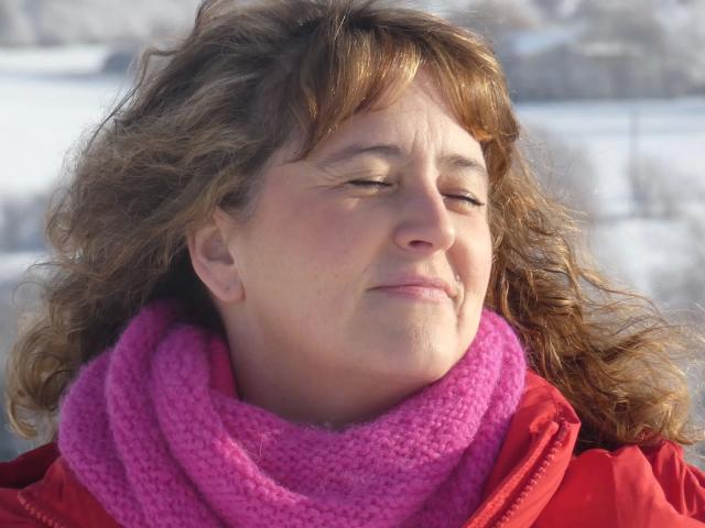 Pamm2023-hot has curly brown hair, closed eyes, and a pink scarf, wearing a red jacket, with a content, slightly smug expression. Snowy background is blurred.