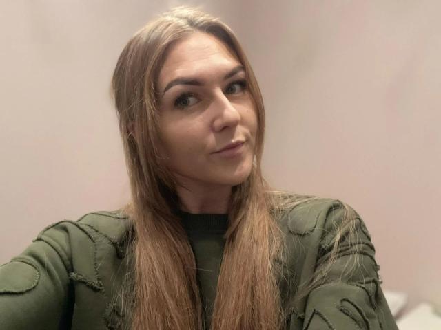GanzNettePaola-hot hat langes, braunes Haar, grüne Augen und ein leicht schmollendes Gesicht. Sie trägt eine grüne, gesteppte Jacke, hat dezentes Make-up und ein leichtes Lächeln..