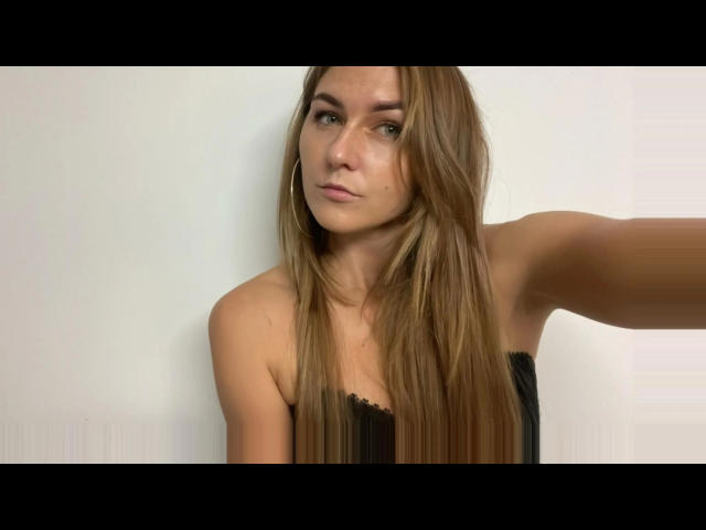 GanzNettePaola – attraktiv mit langen braunen Haaren, grünen Augen und Reifenohrringen – trägt ein schwarzes Top ohne Träger, hat einen leichten Schmollmund und einen erhobenen Arm und blickt direkt in die Kamera..