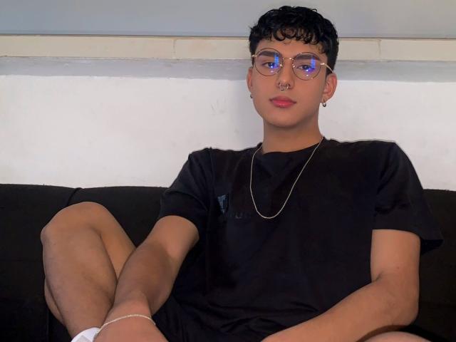 LeJaxon with curly black hair, glasses, nose ring, red lipstick, black t-shirt, and chain necklace, sitting with legs apart on a black couch, wearing white socks.
