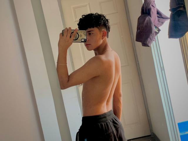 LeJaxon, shirtless, with curly black hair, holding a phone in front of a mirror, wearing black pants, showing a toned back and butt, standing in a sunlit room.