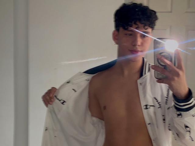 LeJaxon, zonder shirt, met een witte jas, die een foto van zichzelf maakt in een spiegel met een telefoon. Flits van de telefoon, LeJaxon's hand op de jas, zwart krullend haar, licht open lippen..