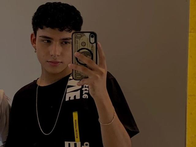 LeJaxon with curly black hair, nose ring, and silver chain, taking a mirror selfie with a phone, wearing a black t-shirt, in a dimly lit room.