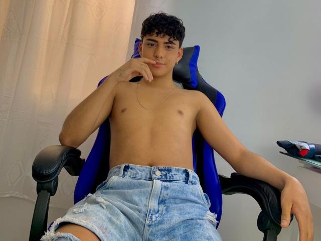 LeJaxon with curly black hair, tan skin, and a nose ring, shirtless, wearing ripped blue denim shorts, sitting in a blue gaming chair, hand resting on his chin.