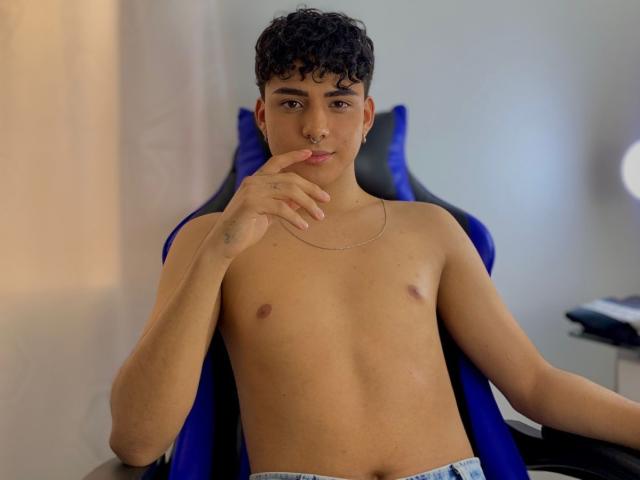 LeJaxon, sin camisa y con cabello negro rizado, está sentado en una silla azul, con un dedo en los labios, tiene un piercing en la nariz, piel clara, pezones pequeños, un collar, pantalones azules ligeramente visibles y una expresión tranquila..
